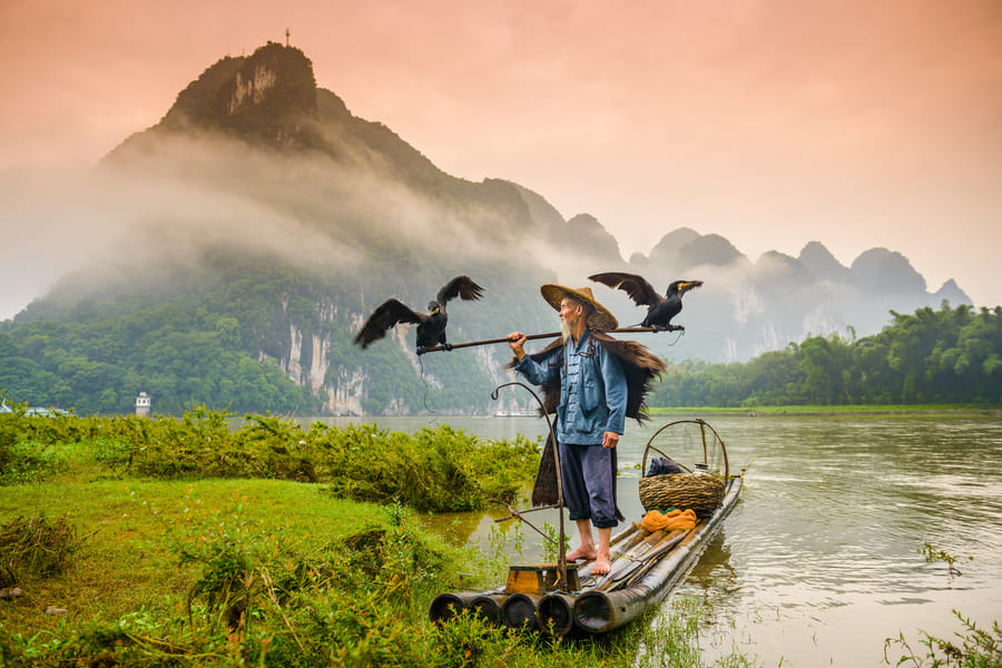 Pescador de cormoran en una lancha en Yangshuo, con montañas de fondo.