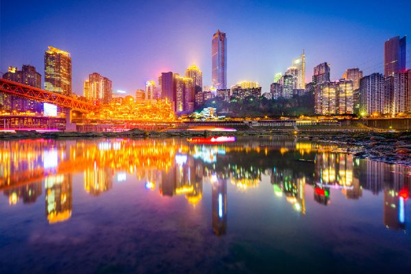 Paisaje de una ciudad de Chongqing donde se aprecia un lago y edificios iluminados al fondo que se reflejan en el lago, la postal es de noche.