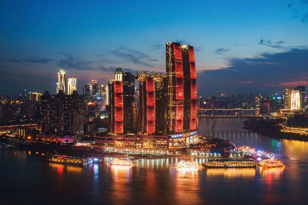 Fotografía de la ciudad del Chongqing en in anochecer con edificios altos iluminados y barcos alrededor.