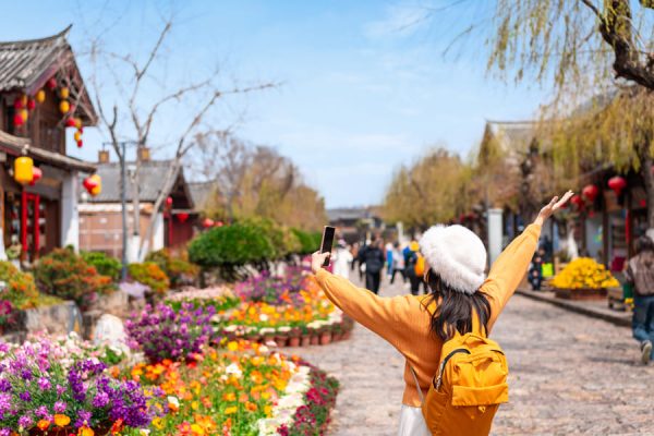 Mujer en una calle de Yunnan con los brazos abiertos y la calle llena de flores de diferentes colores con gente al fondo.